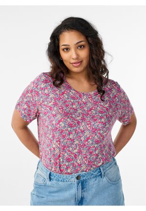 Woman with curly hair wearing a pink floral short-sleeve top and light blue jeans, standing with hands behind her back.
