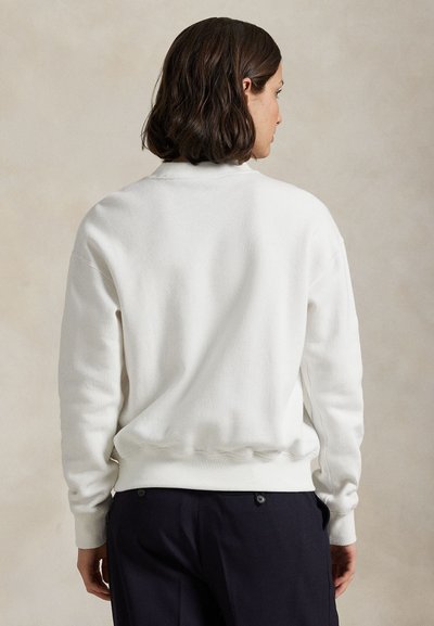 Γυναίκα με κοντά σκουρόχρωμα μαλλιά, που φοράει απλό λευκό πουλόβερ μακρυμάνικο και σκούρο παντελόνι, γυρισμένη πλάτη μπροστά σε ουδέτερο φόντο.