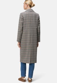 Geruite lange jas in bruin, blauw en crème. Heeft een opstaande kraag, een relaxed fit en een split aan de achterkant. Gecombineerd met blauwe jeans en bruine schoenen.