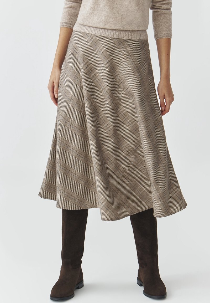 Femme portant une jupe midi trapèze à carreaux beige avec un pull beige et des bottes hautes marron foncé.