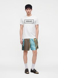 Homme portant un t-shirt blanc Versace, un short à motifs multicolores, des chaussettes blanches et des mocassins noirs avec des touches dorées, debout devant un fond uni.