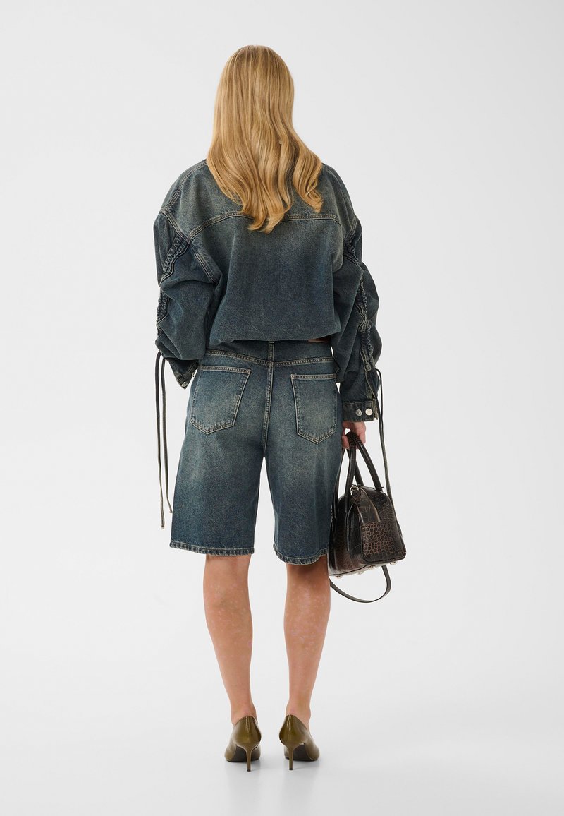 Femme aux longs cheveux blonds portant une veste en jean délavée et un short bermuda assorti, tenant un sac à main texturé foncé et portant des talons verts.