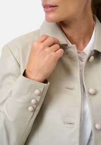 Hellgraue Lederjacke mit kragenförmigem Nacken und drei großen, glatten Knöpfen an den Ärmelabschlüssen. Verfügt über dezente Nähdetails.