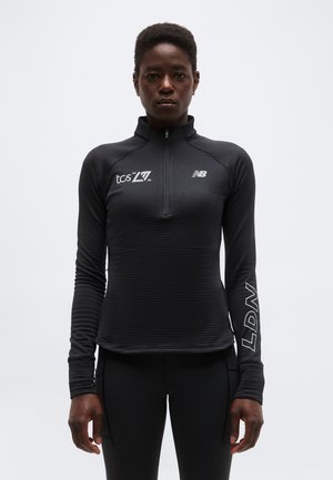 Αθλητικό άτομο που φοράει μαύρο μακρυμάνικο φόρεμα με φερμουάρ και λογότυπα "tcs 2026", "NB" και "LDN", στέκεται μπροστά σε απλό φωτεινό φόντο.