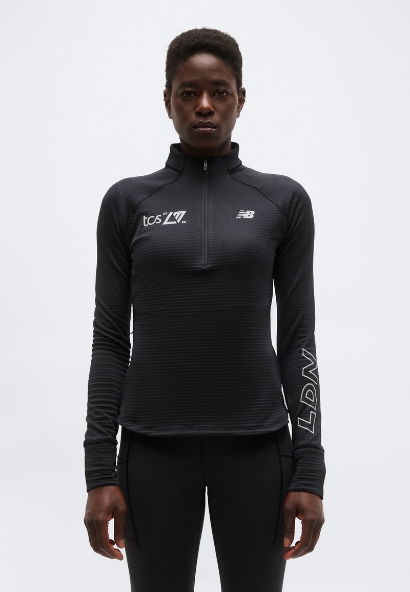 Atletska oseba, oblečena v črno dolgoročno jopico z zadrgo, na kateri so logotipi "tcs 2026", "NB" in "LDN", stoji ob preprostem svetlem ozadju.