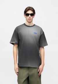 Μπλουζάκι με διαβάθμιση μαύρου χρώματος και κοντά μανίκια, με μπλε λογότυπο στο στήθος. Φορεμένο με χακί παντελόνι cargo.