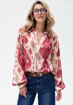 Seidene Hemdbluse mit einem rosa und beige Ikat-Muster, im entspannten Schnitt, mit langen Ärmeln und klassischem Kragen. Kombiniert mit blauen Jeans.