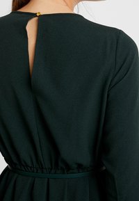 Mörkgrön klänning med långa ärmar, med en nyckelhål baktill och ett elastiskt midjeband med ett matchande bälte. Slät tygtextur.