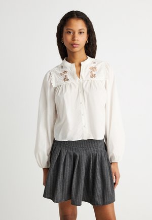 Naine, kes kannab valget pluusi pitskraega ja pikkade varrukatega, tumesiniste triibuliste volditud seelikuga, seisab ühevärvilise tausta ees.