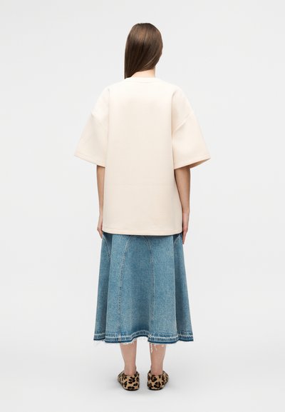 Φαρδιά μπλούζα σε ανοιχτό μπεζ βαμβακερό ύφασμα με κοντά μανίκια, συνδυασμένη με μπλε denim φούστα μήκους μέχρι το γόνατο, με εμφανείς ραφές και ξεφτισμένο τελείωμα.