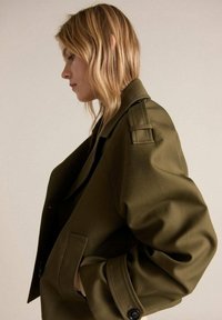 Olive green oversized jacket with a structured design, featuring large lapels, shoulder epaulets, and a relaxed fit. Smooth texture with buttons.