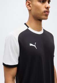 Junger Mann trägt ein schwarz-weißes Puma-Sportshirt mit Puma-Logo auf der Brust, schaut nach rechts vor neutralem hellem Hintergrund.