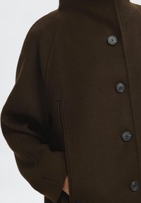 Manteau en laine marron avec un col haut, comportant de grands boutons noirs et des poches latérales. Le tissu a une finition douce et texturée.