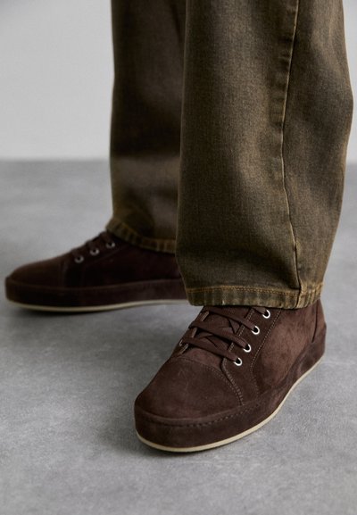 Chaussures en daim marron avec un design bas, un laçage à l'avant et une semelle en caoutchouc de couleur claire, présentées avec un pantalon vert olive.