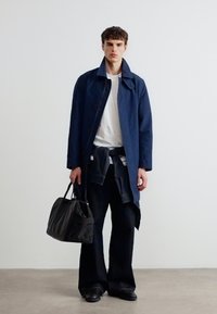 Νεαρός άνδρας με μπλε παλτό, λευκό μπλουζάκι, μαύρο παντελόνι, κρατώντας μαύρη τσάντα, στέκεται μπροστά σε λευκό τοίχο και σκυρόδεμα δαπέδου.