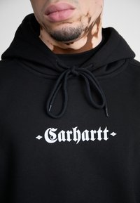 Black hoodie made of cotton, featuring a drawstring hood, front pocket, and white "Carhartt" logo printed on the chest.