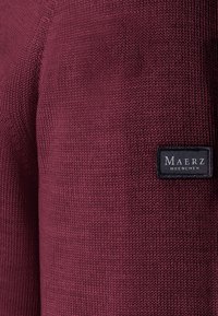 Burgundský pletený svetr s texturovaným povrchem, na kterém je černá nášivka s bílým textem "Maerz Muenchen."