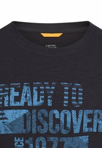 Marineblå bomulds T-shirt med rund halsudskæring, med fed blå grafisk tekst "KLAR TIL AT OPDAGE" og en lille gul accent-tag inde i halsudskæringen.