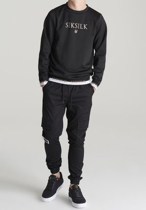 Homme portant un sweat-shirt noir SikSilk, un pantalon cargo noir et des baskets noires à semelles blanches, debout les mains dans les poches.