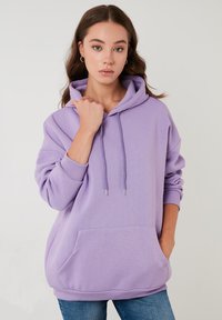 Sudadera de algodón morada con un bolsillo frontal, cordones ajustables y puños acanalados. El modelo la lleva sobre unos jeans azules, contra un fondo liso.