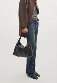 Zwarte leren handtas met een slouchy ontwerp, brede schouderband en subtiele hardwaredetails, vastgehouden door een persoon in een bruine jas en blauwe jeans.