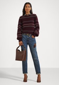 Bordeaux strikkegenser med mønstrede striper, blå denimjeans med detaljerte lapper, og en brun skinn totebag i hånden.