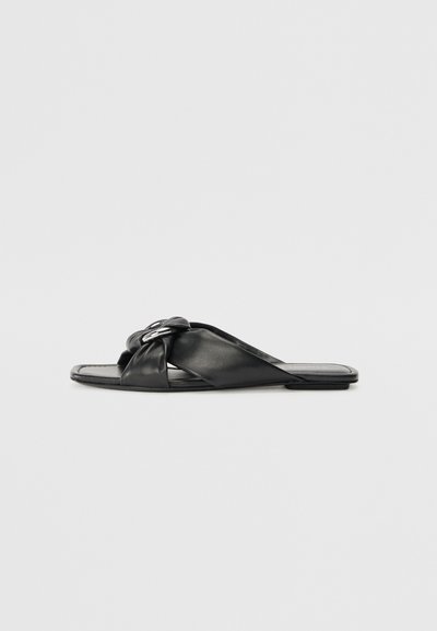Sandales en cuir noir avec un design noué, semelle plate et style minimaliste. Texture lisse, sans embellissements ni quincaillerie visibles.