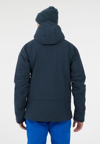 Marineblauwe waterdichte jas met een capuchon, gestructureerde stof, verstelbare manchetten en een ontspannen pasvorm, gedragen met blauwe broek.
