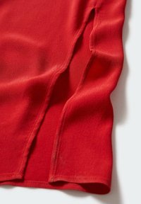 Top rouge avec une texture lisse, présentant des fentes sur les côtés et un ourlet légèrement incurvé. Le tissu tombe doucement, mettant en valeur son design léger.