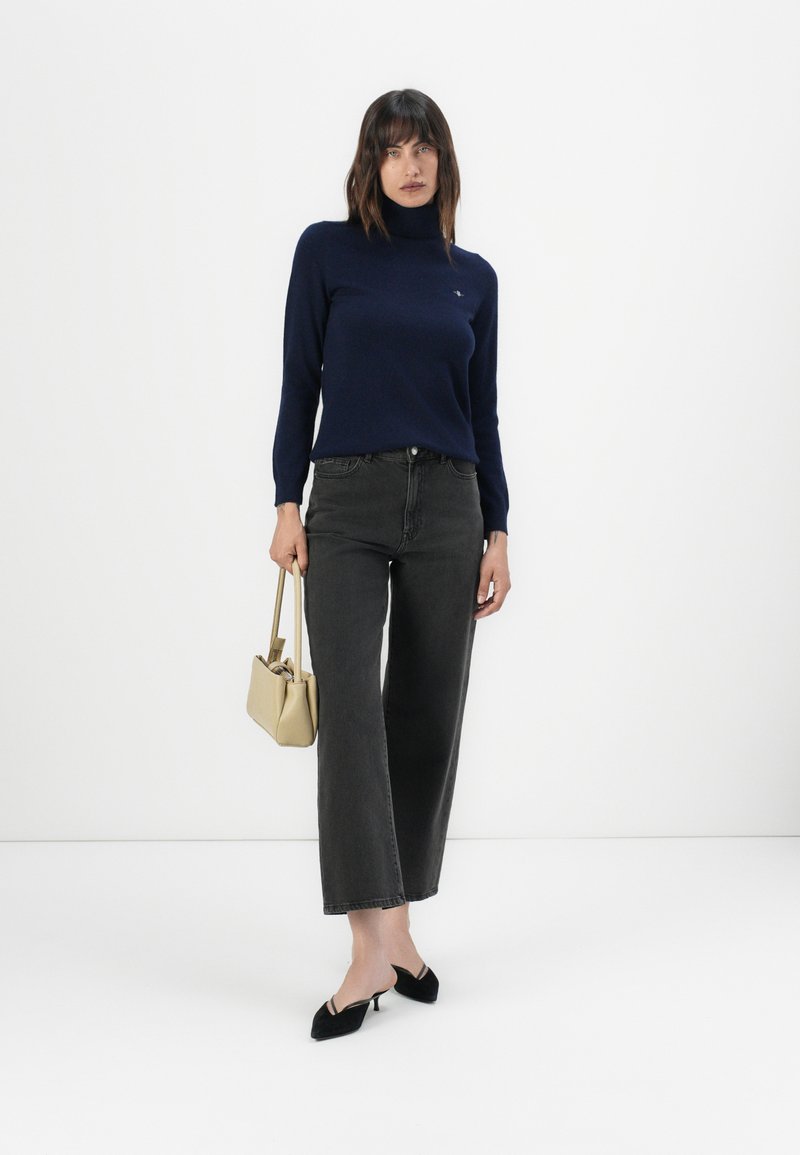 Eine Frau trägt einen marineblauen Rollkragenpullover, schwarze weit geschnittene Jeans, schwarze Kitten-Heels und hält eine beige Handtasche vor einem schlichten weißen Hintergrund.