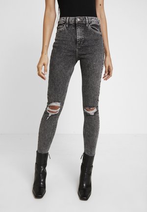 Kvinna som bär högmidjade, svarta acid-wash, slitna skinny jeans med sprickor vid knäna och svarta ankelskor, stående mot vit bakgrund.