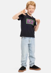 Camiseta negra con un vibrante logo verde y morado, jeans azul claro y zapatos negros con suelas marrones. El niño está posando de manera juguetona.