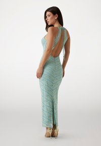 Een lange, mintgroene gebreide jurk met een zigzagpatroon, een open rug en verstelbare bandjes, met een hoge split en stilettohakken.