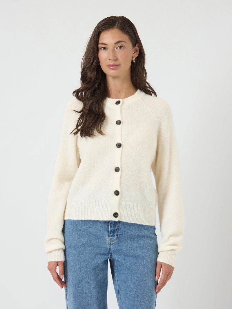 Mulher com cabelo castanho longo, vestindo um cardigã creme com botões e jeans azuis, de pé em frente a um fundo claro e simples.