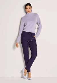 Vrouw in een lavendelkleurige coltrui, donkere paarse joggingbroek met trekkoord en lavendelkleurige hoge hakken, staand tegen een lichte muur.