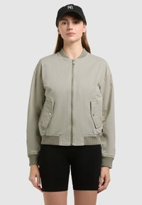 Femme portant une veste bomber gris clair avec des poches frontales, un short noir, et une casquette de baseball noire avec un logo blanc, debout devant un fond uni.