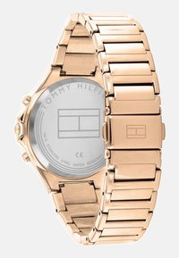 Reloj de acero inoxidable en color oro rosa con pulsera de eslabones, caja redonda y un fondo claro con la marca que muestra "TOMMY HILFIGER". Resistente al agua.