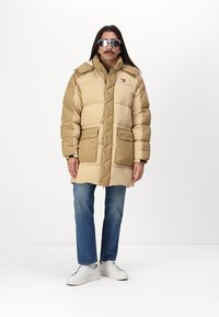 Beige dunsjakke med quiltet design, featuring hette, store frontlommer og merkevarelogo. Kombinert med blå jeans og hvite joggesko.