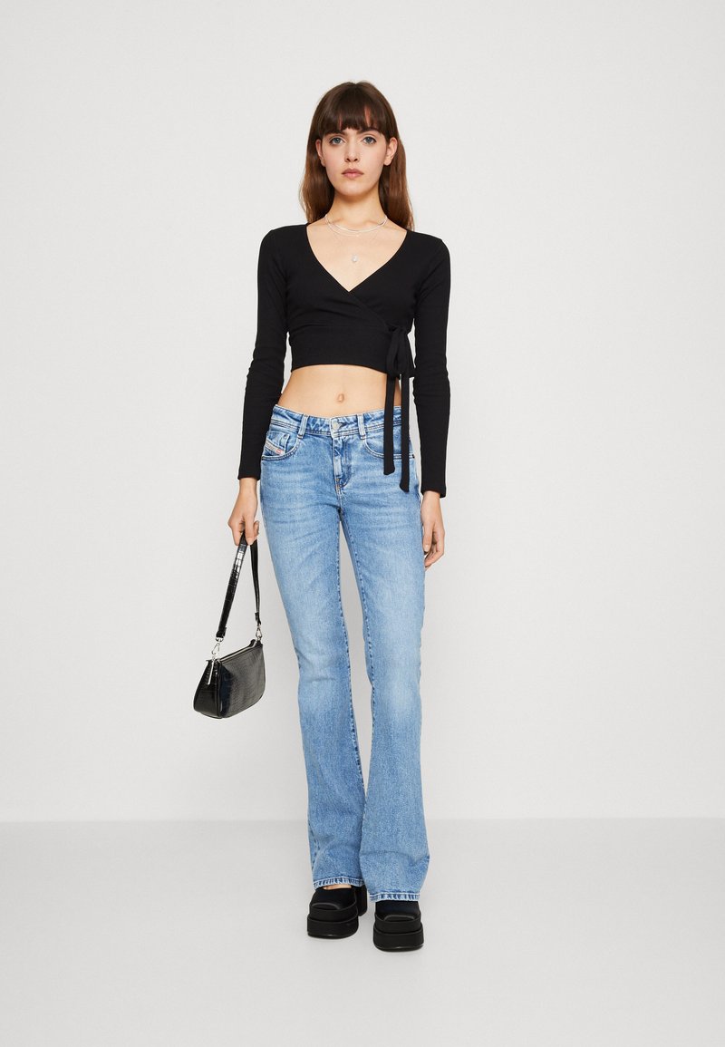 Frau in einem schwarzen langärmligen Crop-Top, hellblauen Schlagjeans, schwarzen Plateauschuhen, die eine kleine schwarze Handtasche vor einem einfarbigen Hintergrund hält.