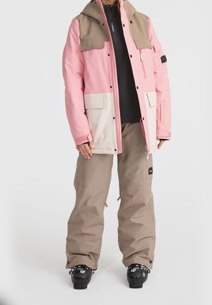 Persoon die een winterjas met kleurvlakken in roze en beige draagt, beige sneeuwbroek, zwarte skischoenen, staand tegen een effen achtergrond.
