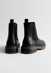 Botas Chelsea de cuero negro con paneles elásticos a los lados, punta redondeada y suela de goma marrón gruesa. Cuenta con una pestaña para tirar en la parte trasera.