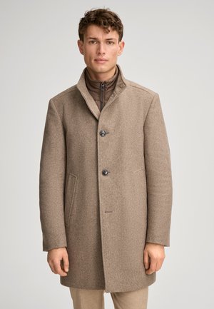 Mann trägt einen braunen, geknöpften Wollmantel über einer Reißverschlussjacke, steht vor einem schlichten hellgrauen Hintergrund.