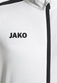 Witte sportjack met zwarte rits en accenten. Voorzien van een gestructureerd zwart logo "JAKO" op de linkerborst, van gladde stof.