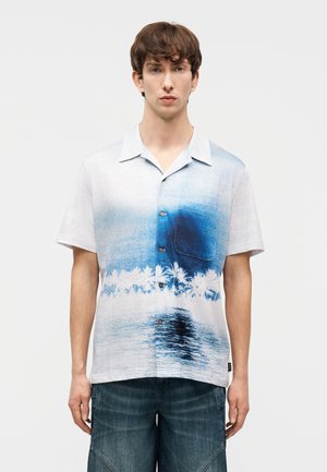 Άνδρας με πουκάμισο με κοντά μανίκια, κουμπωτό, με μπλε και λευκό διαβαθμισμένο χρώμα και εκτύπωση με φοίνικες και παραλία, συνδυασμένο με σκούρα τζιν σορτς.