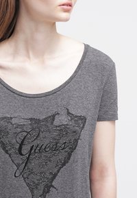Šedé tričko s krátkým rukávem, které má černý krajkový design srdce a text "Guess" spolu s dekorativními prvky. Jemná látka se širokým výstřihem.