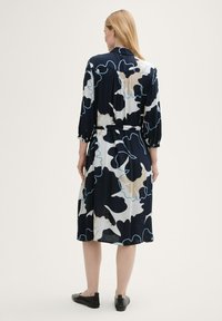 Marineblaues Kleid mit abstraktem Creme- und Blaumuster, Dreiviertelärmel, taillierter Schnitt, knielang, leichtes Material, flache schwarze Schuhe.