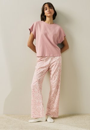 Femme debout contre un mur portant un haut rose ample à manches courtes et un pantalon large rose à motifs avec des chaussettes blanches sur un sol texturé.