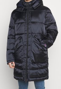 Abrigo acolchado azul marino con cremallera frontal, cuello alto y puños elásticos. Cuenta con dos bolsillos laterales y un detalle de logotipo en la manga.