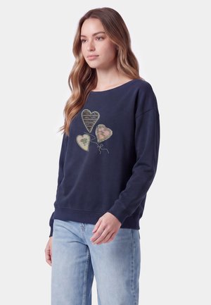 Ung kvinde med langt bølget hår iført en navy blå sweatshirt med tre paliet-hjerte designs og lyseblå jeans, stående foran en ensfarvet baggrund.