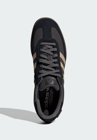 Zapatilla negra con malla y texturas de ante grises, con tres franjas beige, cordones redondos y una plantilla acolchada visible en el interior.